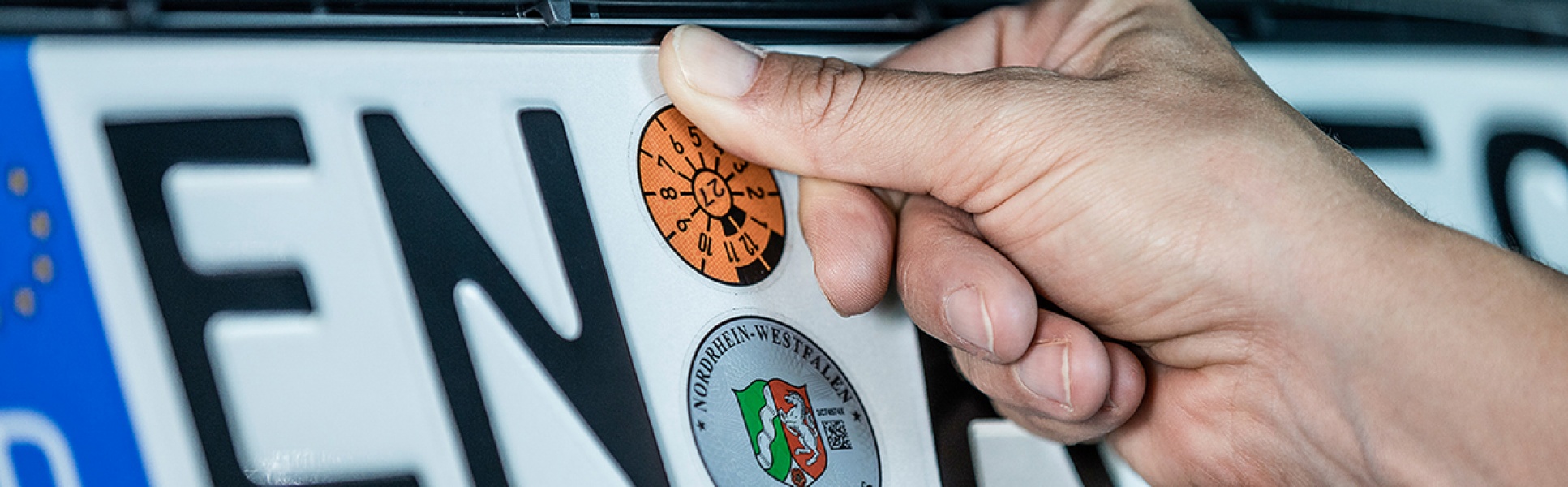 KFZ-Kennzeichen auf die eine Hand eine frische TÜV-Plakette klebt